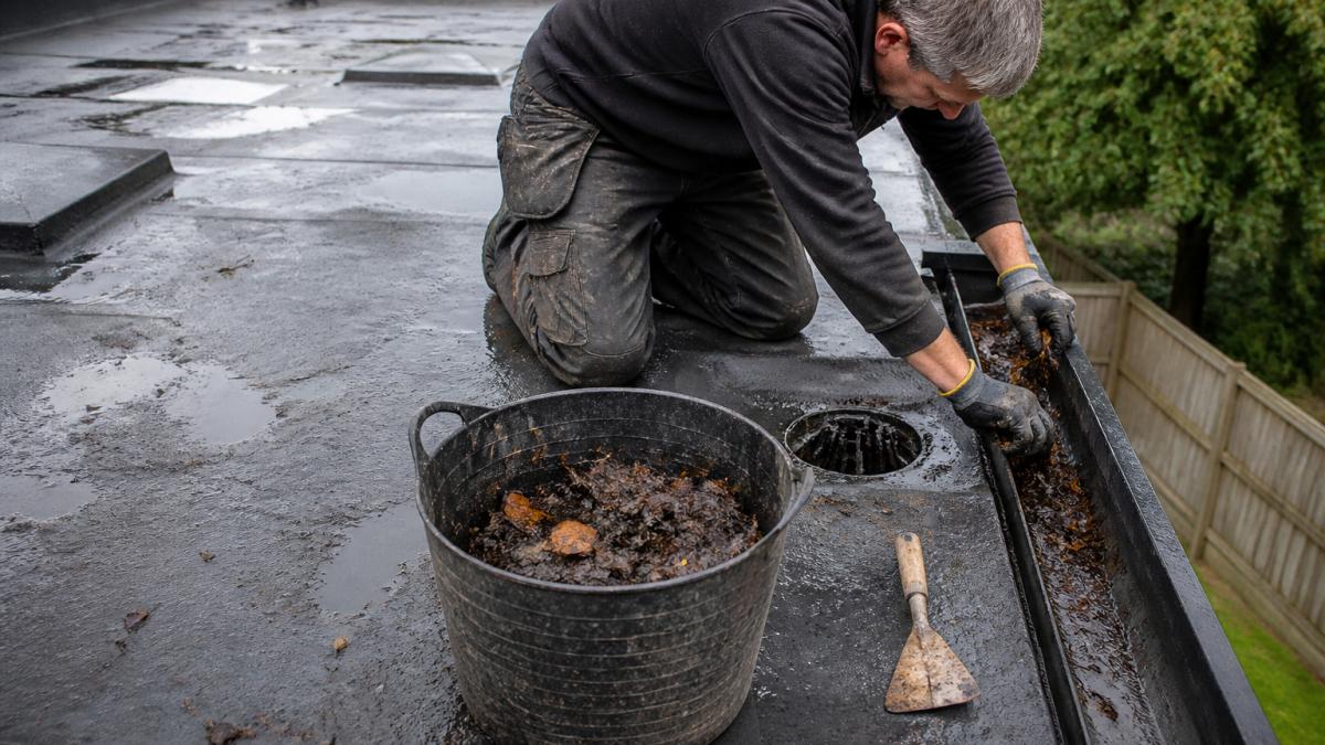 A roofer in work clothes clearing leaves and debris from a flat roof drainage outlet and gutter, with a trowel and bucket nearby on the roof surface