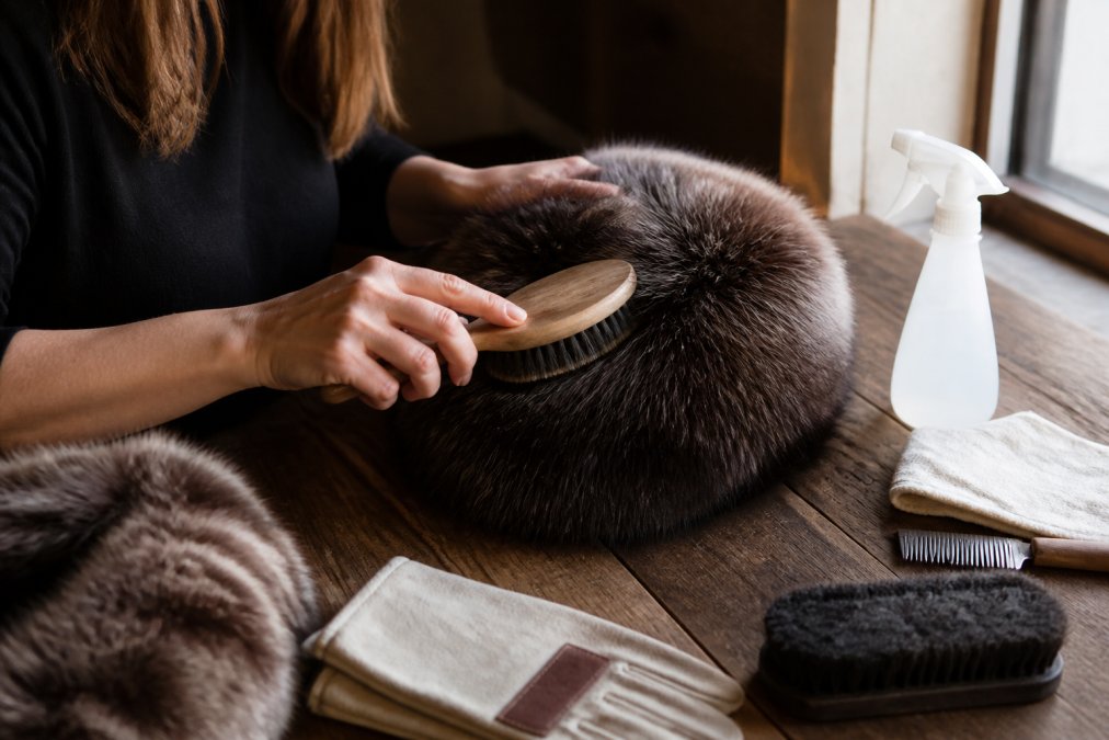 Close-up of a person gently brushing a high-quality fur hat with a soft grooming brush on a wooden surface, warm natural window light illuminating the fur texture