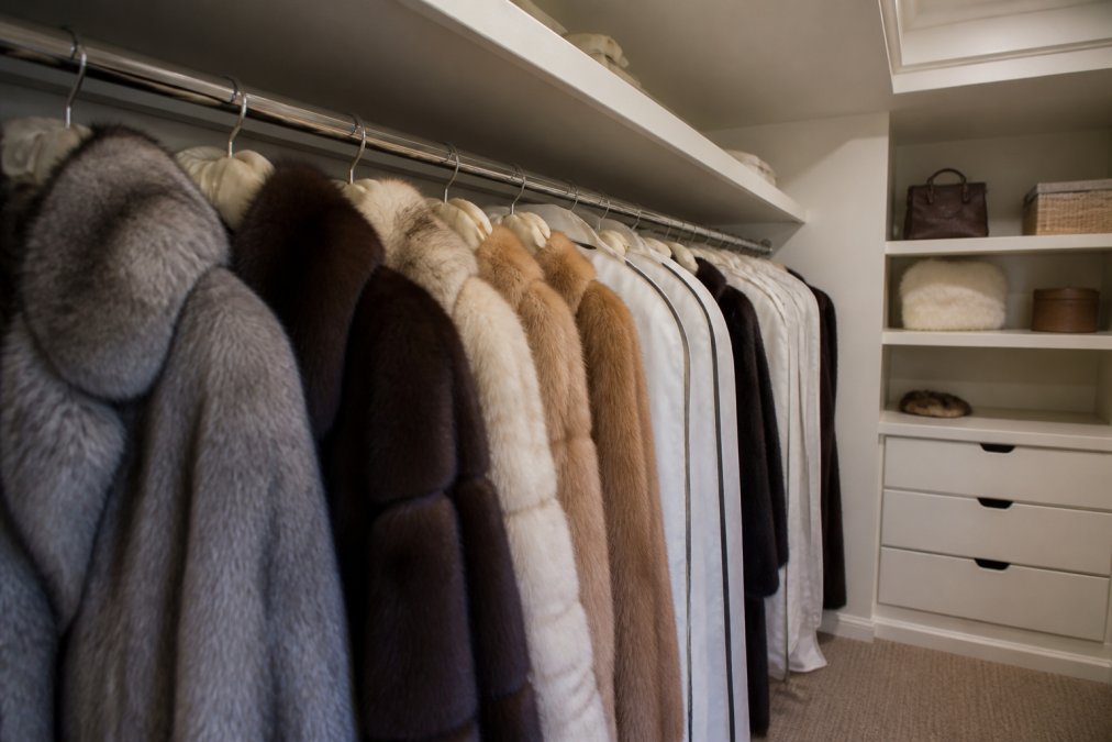 A neatly organized closet with fur coats hanging on wide padded hangers in a cool, well-ventilated space, cloth garment bags visible on several pieces, soft ambient lighting