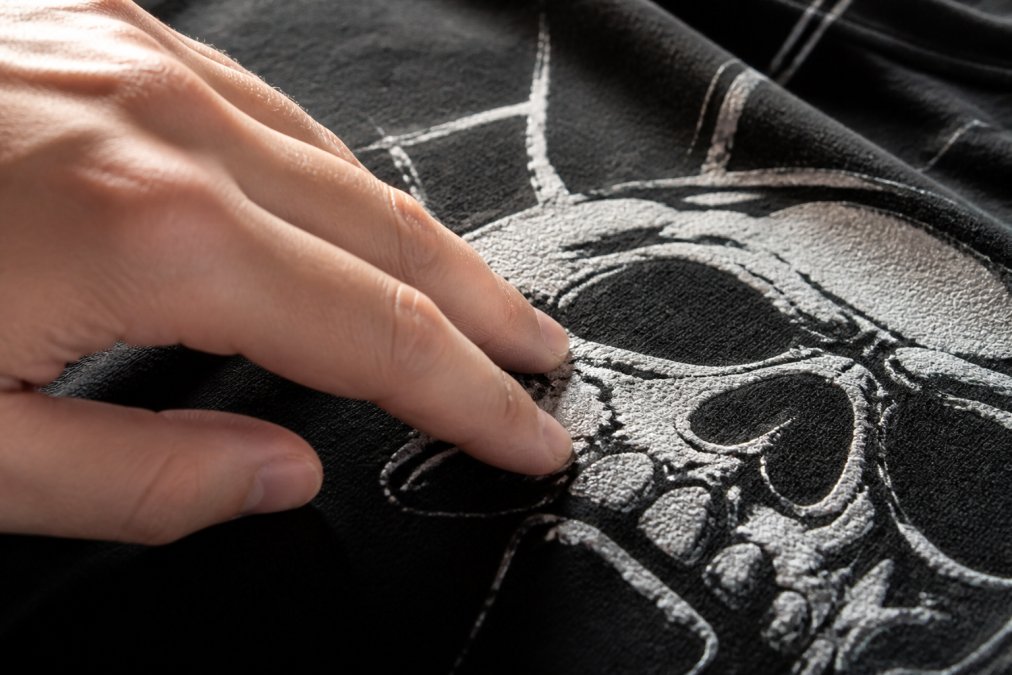 Close-up of a person's fingers rubbing the surface of a black graphic t-shirt to feel the raised texture of a plastisol screen print in natural window light