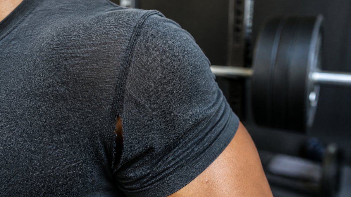 Close-up of a lifter's shoulder and armpit area on a worn gym shirt, showing stretched fabric and a small tear along the seam, with a barbell and weight plates blurred in the background of a dim gym
