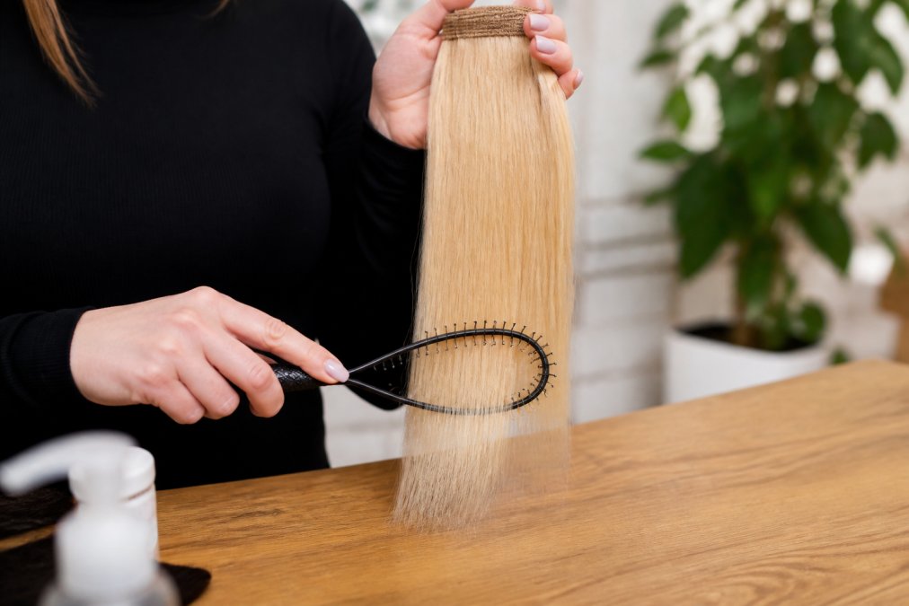 Close-up of a woman's hands using a loop brush on silky blonde hair extensions, showing proper technique with gentle downward strokes on the lower sections