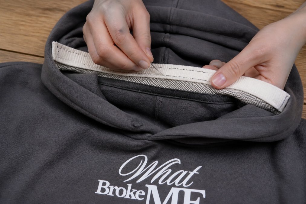 Close-up of a seamstress's hands carefully stitching elastic tape and twill tape onto a hoodie neckline facing with needle and thread, showing detailed reinforcement stitching technique