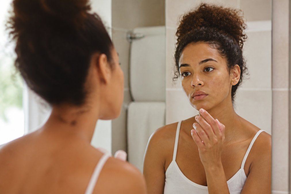 A woman's hand hovering near her face in a mirror reflection, showing restraint from touching a blemish, soft bathroom lighting