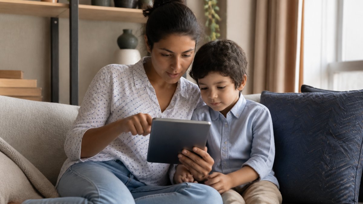 A parent and young child sitting together on a living room sofa, the parent holding a tablet and pointing at the screen while the child watches, warm natural light from a window
