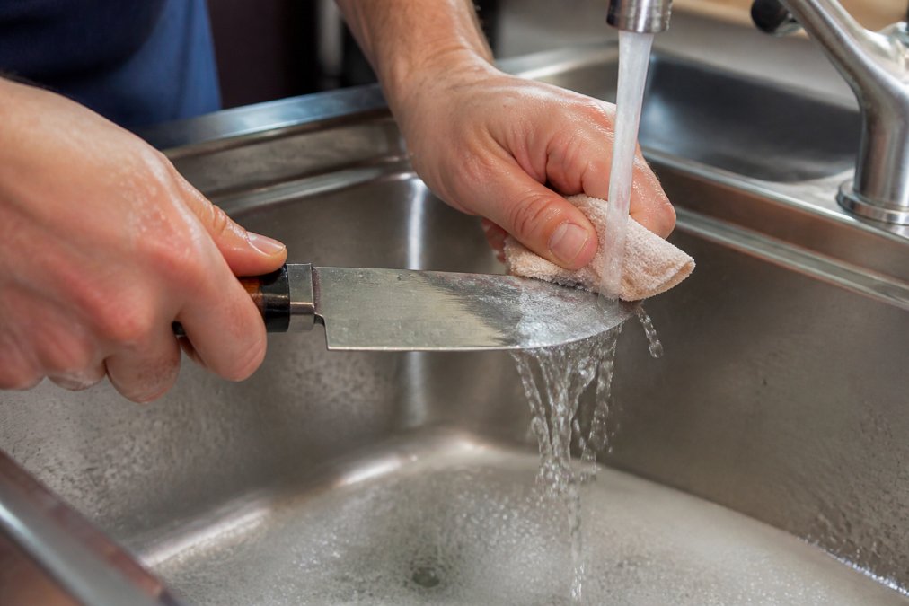 Close-up of hands washing a custom knife blade under running water with a soft cloth, showing proper gentle cleaning technique with warm soapy water and careful handling