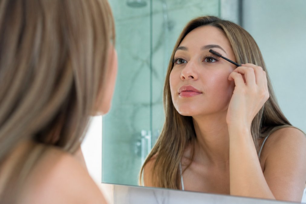 A close-up of a person gently brushing their eyelash extensions with a clean spoolie brush in a bright bathroom mirror, showing the proper aftercare technique
