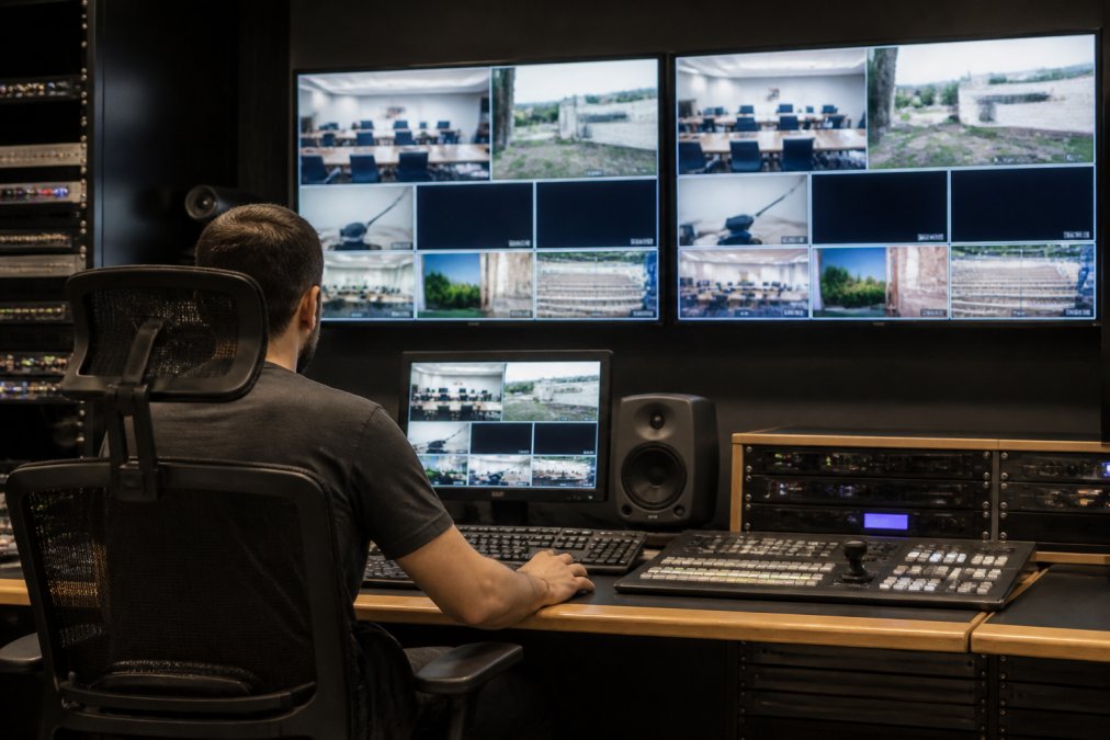 A broadcast engineer seated at a professional webcasting control room desk, monitoring multiple large screens displaying stream health dashboards and live video feeds, with rack-mounted hardware and indicator lights visible in the background