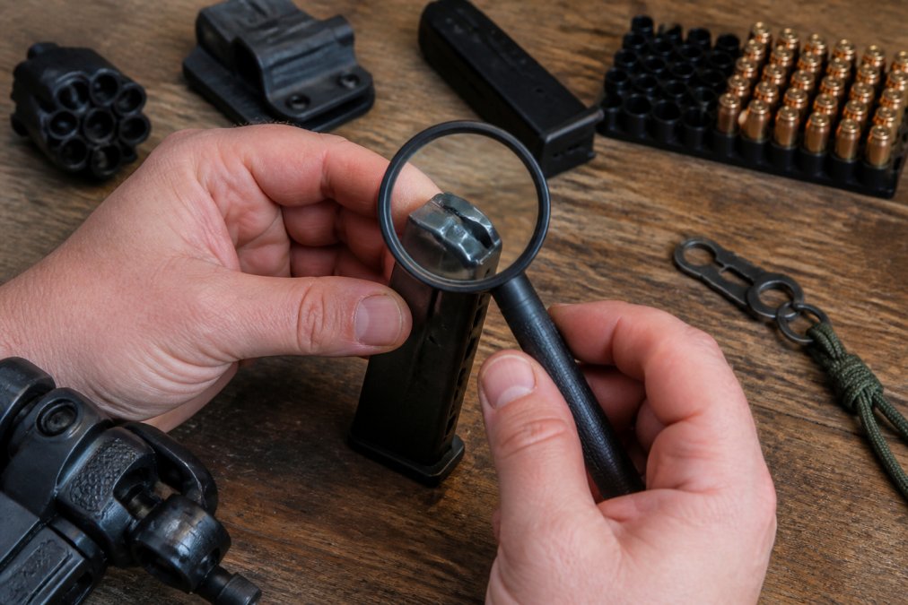 Close-up of hands holding a 9mm magazine under bright overhead lighting, examining the feed lips with a magnifying glass to inspect for visible cracks, deformation, and wear marks