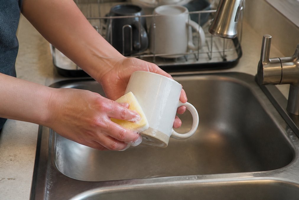 Close-up of hands carefully hand-washing a ceramic mug with a soft sponge in a stainless steel sink, with a dish rack nearby