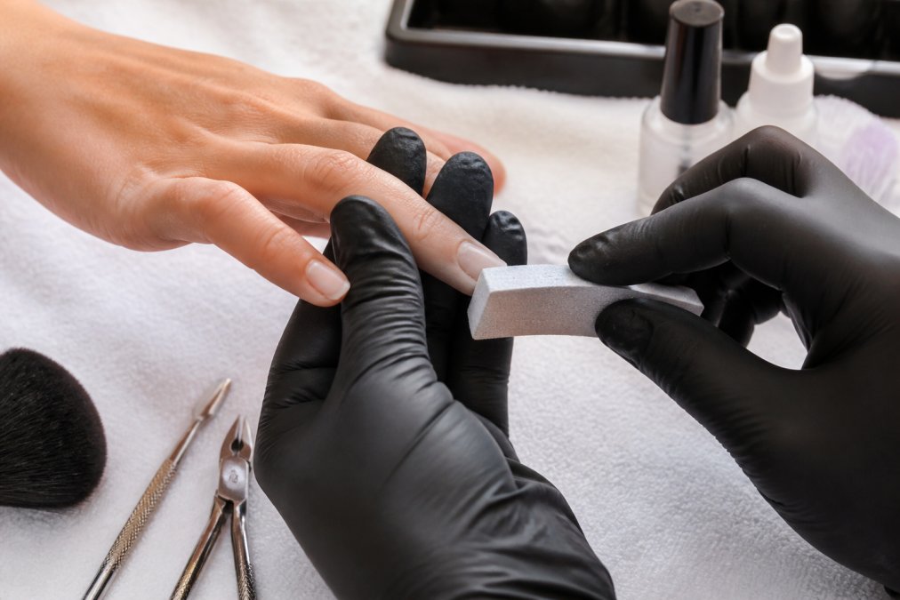 Close-up of a nail technician's hands using a fine-grit buffer on a client's natural nail plate, with prep tools and primer bottle visible on a clean salon towel