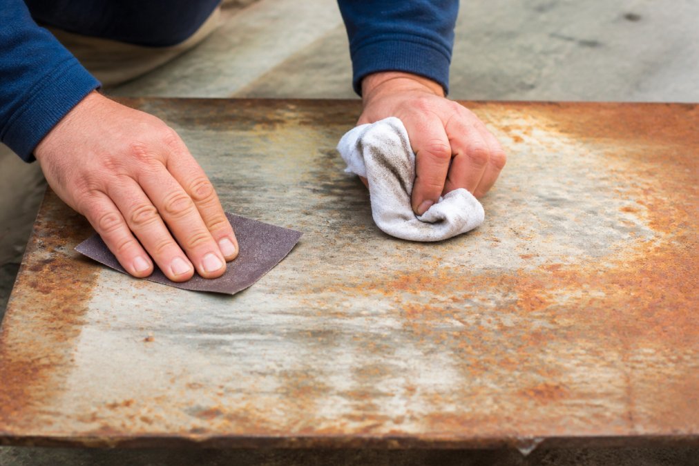 Close-up of hands using sandpaper and degreasing cloth on a weathered metal sign surface, showing texture preparation and cleaning work in natural daylight