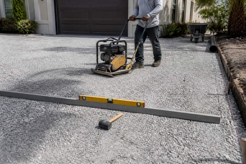 How to Prevent Paver Sinking: A Base Prep Guide 2 Worker using a plate compactor on crushed stone to prevent paver sinking on a residential driveway project.