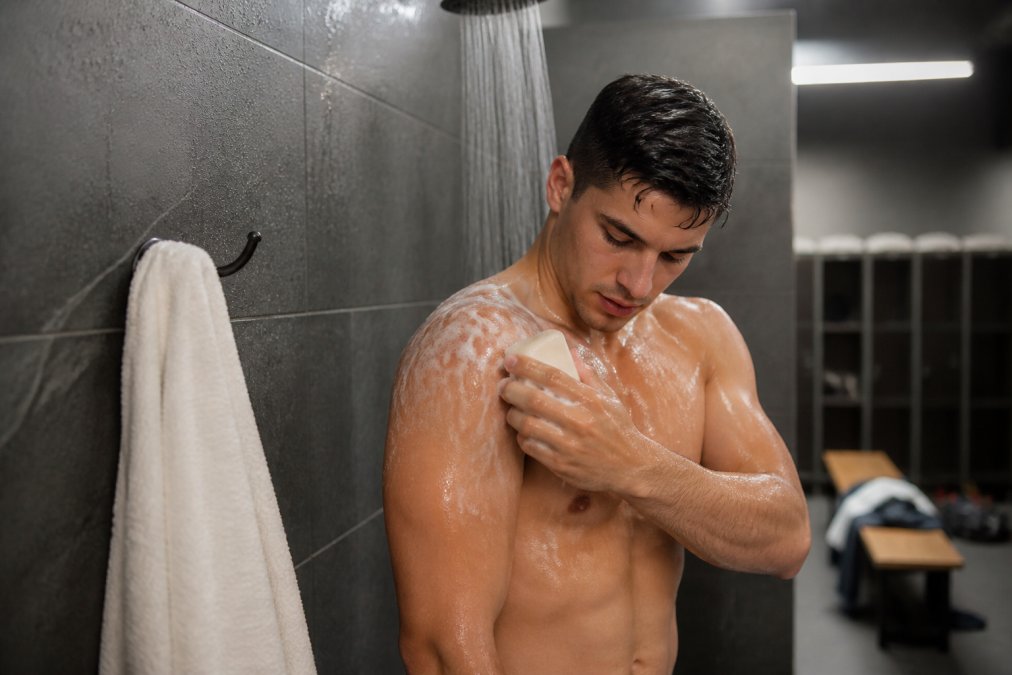 A BJJ athlete in a gym locker room showering with a bar of handmade soap, water running, clean towel nearby, realistic candid scene