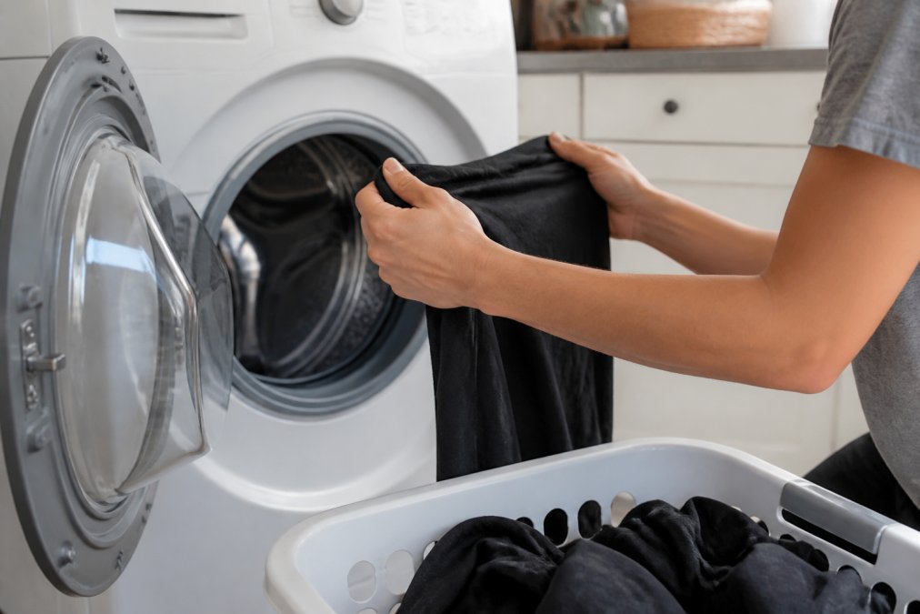 Close-up of hands turning a dark-colored t-shirt inside out before placing it in a front-loading washing machine, print facing inward, bright laundry room lighting