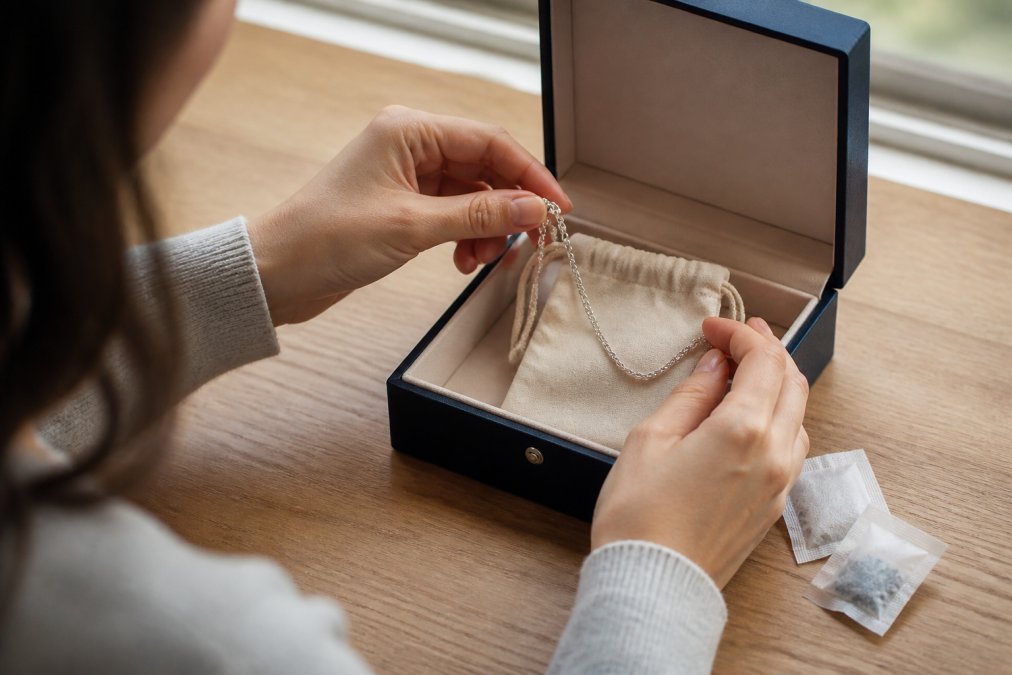 Close-up of hands placing a sterling silver chain into a soft anti-tarnish pouch inside a lined jewellery box, with small silica gel packets visible beside it on a light wooden surface