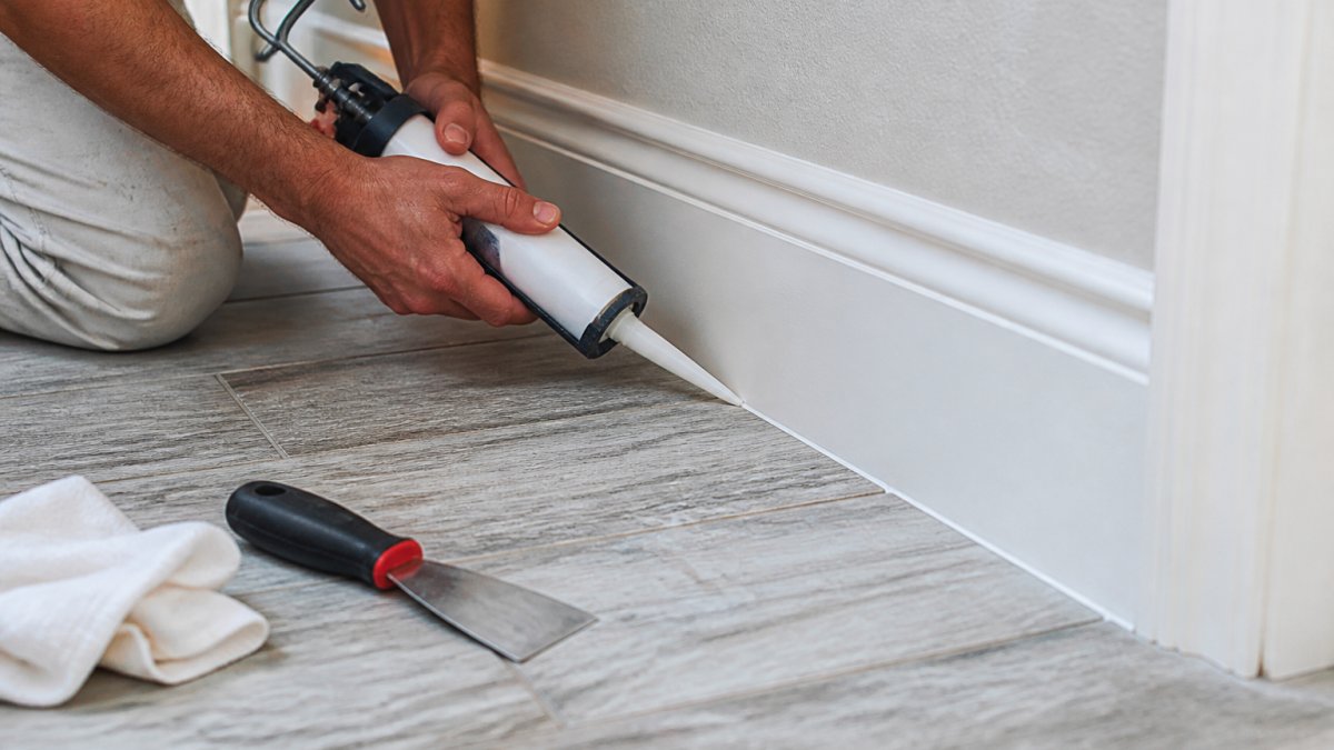 Close-up of hands applying caulk along a baseboard gap where a wall meets the floor in a bright Florida home, with a caulking gun and putty knife nearby