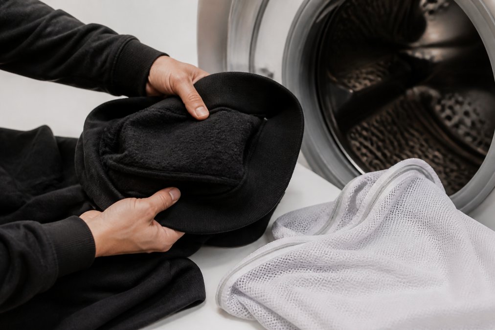 Close-up of hands turning a dark heavyweight cotton hoodie inside out before placing it into a washing machine, laundry mesh bag visible nearby on a clean white surface