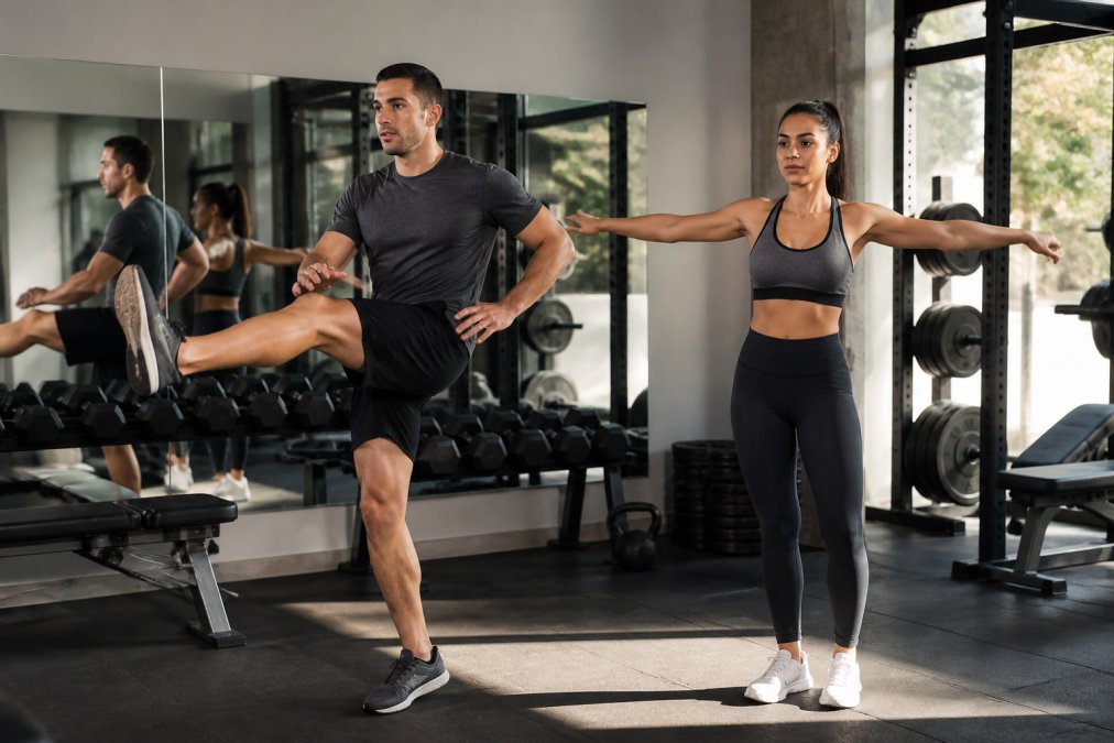 Athlete performing dynamic stretching movements before strength training session, doing leg swings and arm circles in a bright gym setting with mirrors