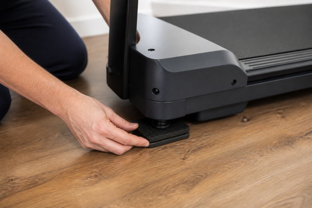 Close-up of hands placing rubber isolation pads under each corner of a treadmill on a wooden apartment floor, showing proper positioning with pad visible between treadmill foot and floor