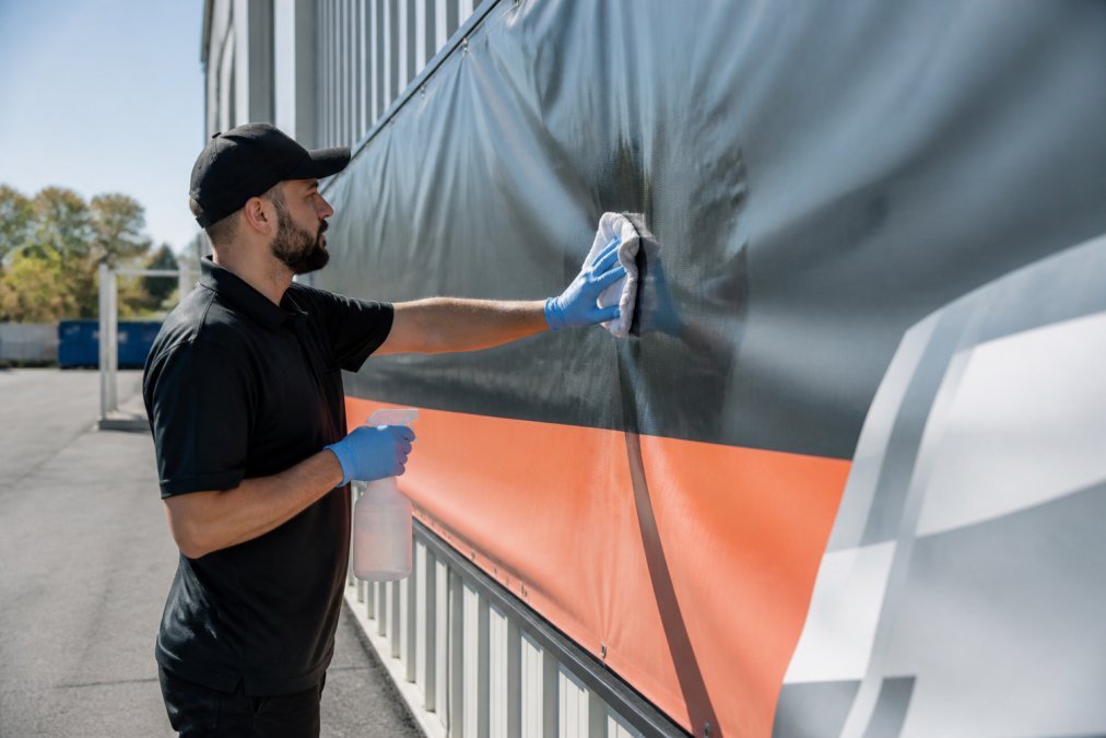 A sign shop worker wearing gloves carefully cleaning a large outdoor vinyl banner with a soft cloth and spray bottle, bright daylight, professional setting