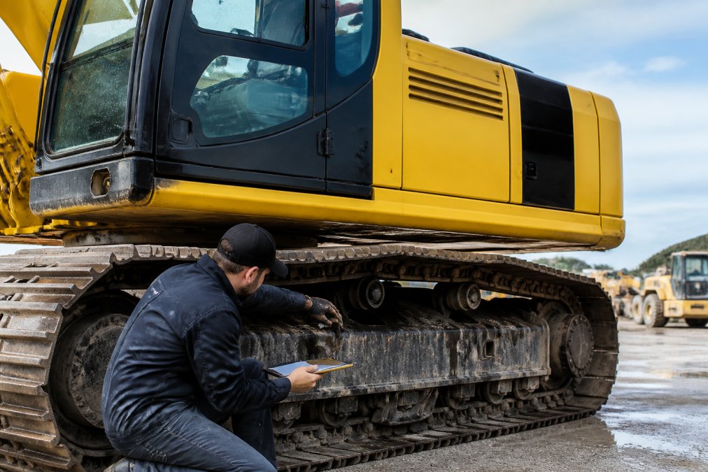 Heavy excavator being cleaned and inspected before sale, technician checking undercarriage components and documenting condition with tablet