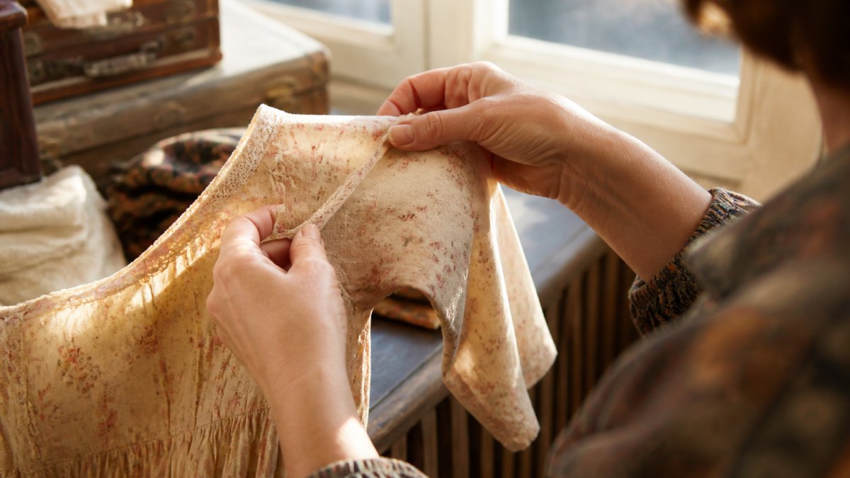 Close-up of hands holding vintage dress fabric, examining seams and weave texture under natural window light to assess condition