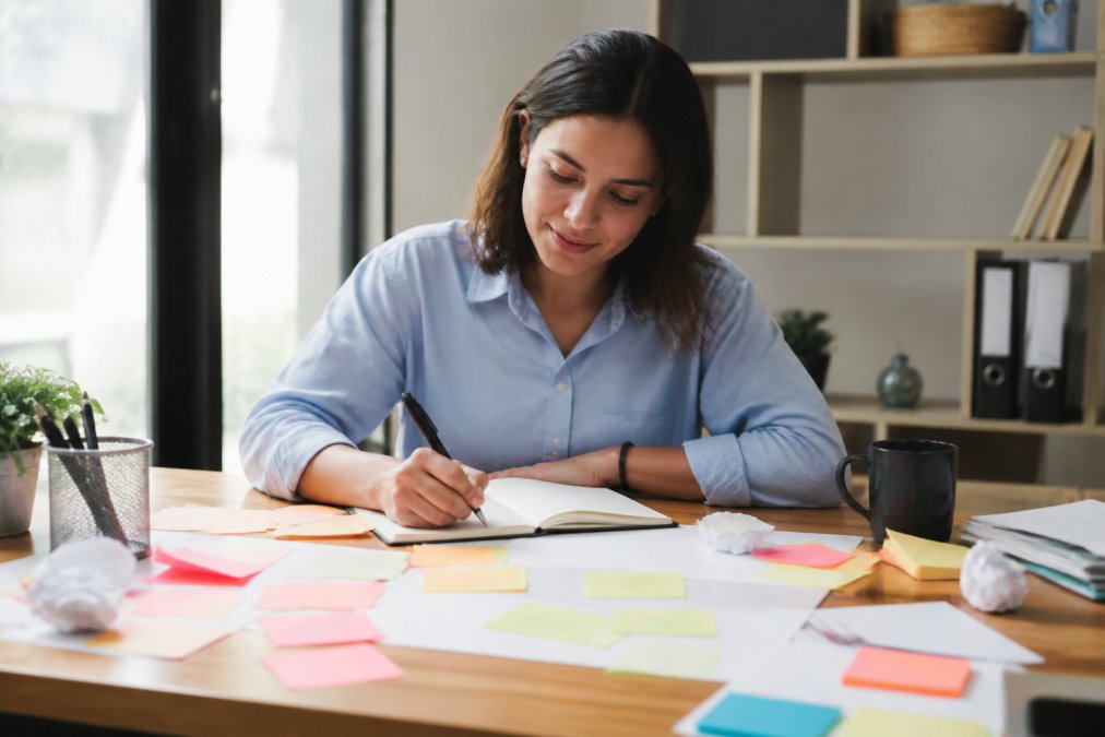 Professional sitting at desk with notebook and pen, writing down tasks from scattered sticky notes and papers, looking focused and relieved as ideas flow onto the page