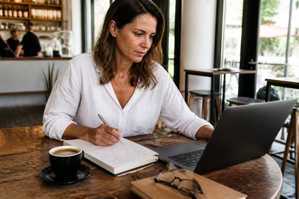 Consultant sitting at desk reviewing time-tracking data on laptop screen, notebook with process notes beside coffee cup, focused on identifying patterns in work schedule
