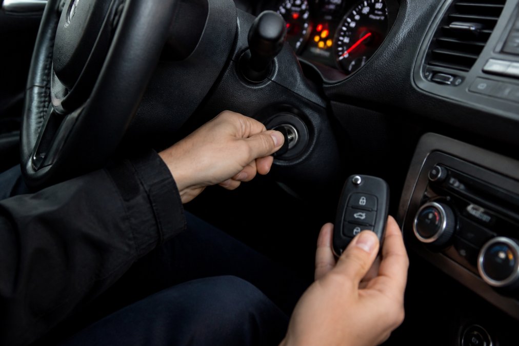 Close-up of a person's hands inserting a car key into the ignition cylinder while holding a key fob remote in the other hand, inside a dimly lit vehicle interior with dashboard lights visible