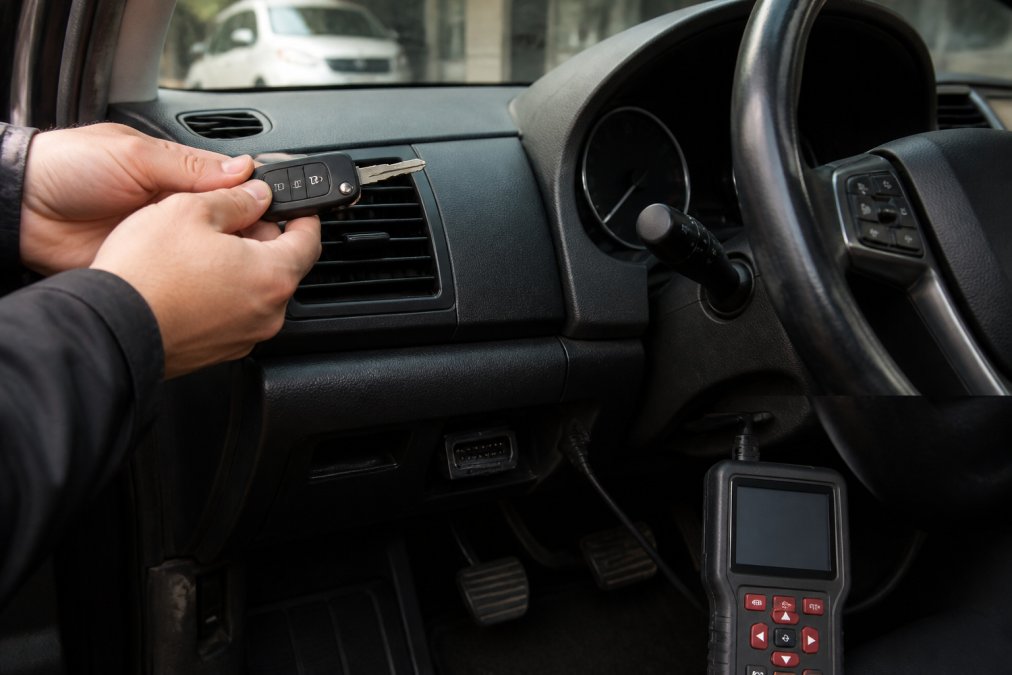 Hands holding a car key against a vehicle's dashboard with VIN visible on the windscreen, OBD-II diagnostic port visible beneath the steering column with a scanner device nearby