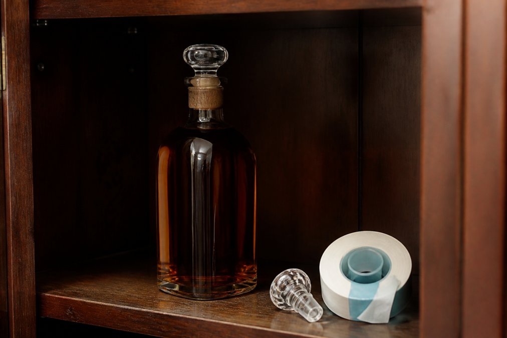 A close-up of a bourbon bottle standing upright inside a dark wooden cabinet, with a glass stopper and a roll of parafilm on the shelf beside it