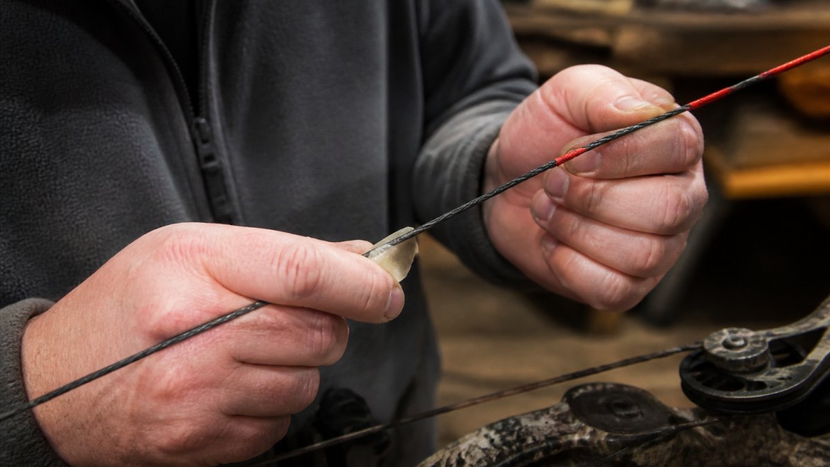 Archer's hands applying wax to bowstring strands with steady friction motion, showing wax distribution along exposed fibers in natural workshop lighting