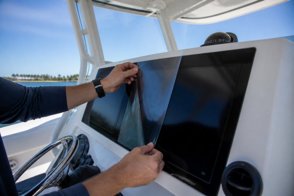 Close-up of hands applying a clear screen protector film to a large marine GPS display unit mounted on a center console helm, with the boat's T-top frame visible in the background under bright sunlight