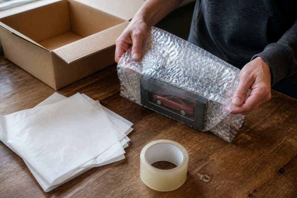 A collector's hands wrapping a diecast car box in a layer of bubble wrap at a clean table, with acid-free tissue paper, tape, and a corrugated box nearby