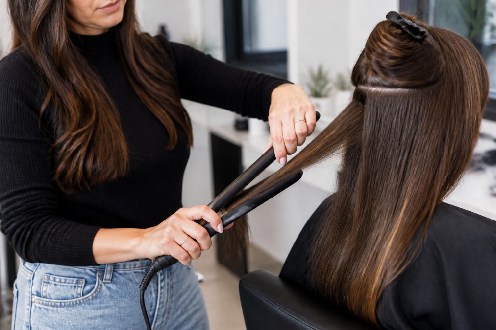 Professional demonstrating proper flat iron technique on a section of long hair, showing correct hand positioning and single-pass motion through a strand in natural lighting