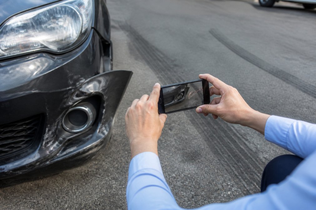 Person's hands documenting vehicle damage with a smartphone camera at crash scene, with damaged bumper and skid marks visible on pavement in background, daylight