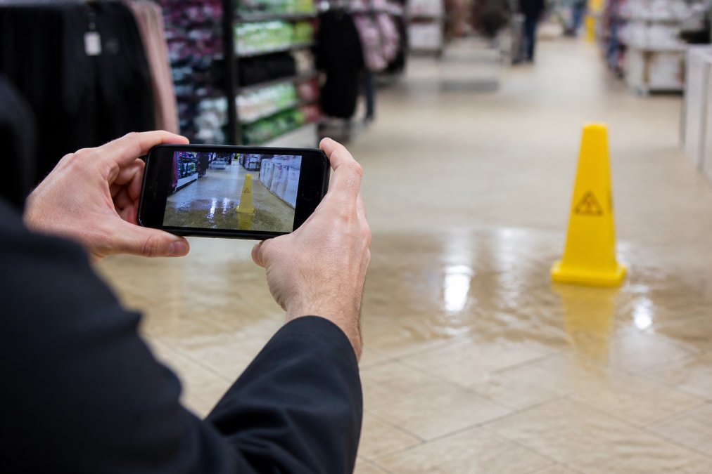 Close-up of a smartphone capturing detailed photos of a wet floor marked with a yellow caution cone in a retail store, documenting the hazardous condition