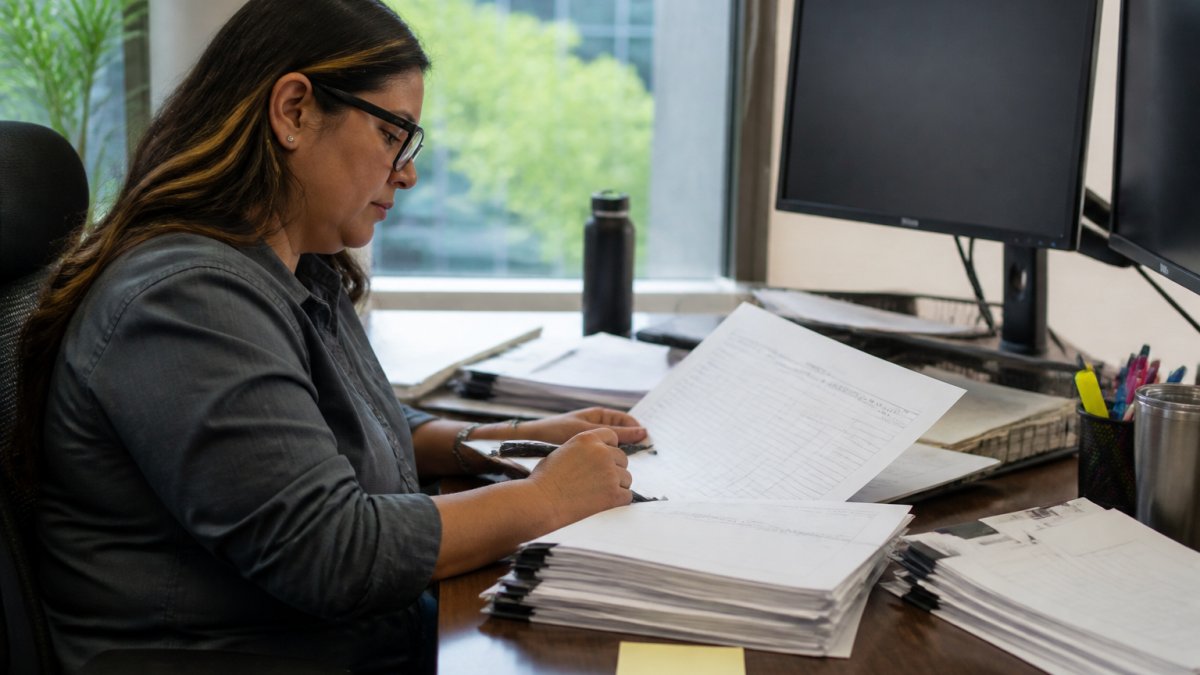 Business development manager at desk reviewing printed bid documents and making notes, with multiple monitors displaying government procurement portals in the background, natural office lighting