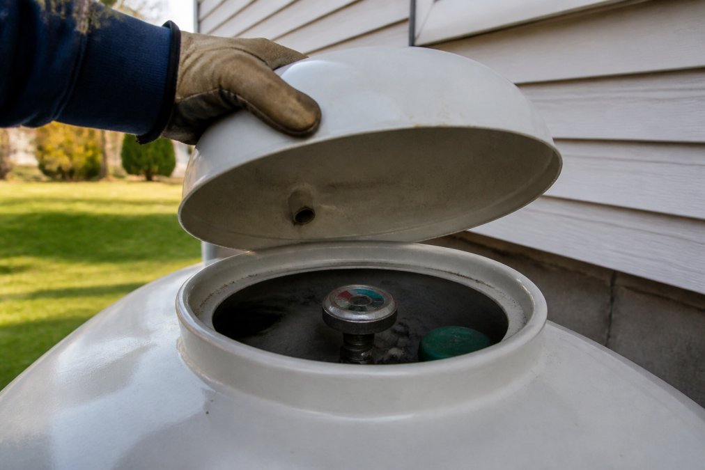 Close-up of a white propane tank dome lid being lifted open by a gloved hand to reveal the float gauge dial underneath, outdoors next to the side of a residential home on a clear afternoon