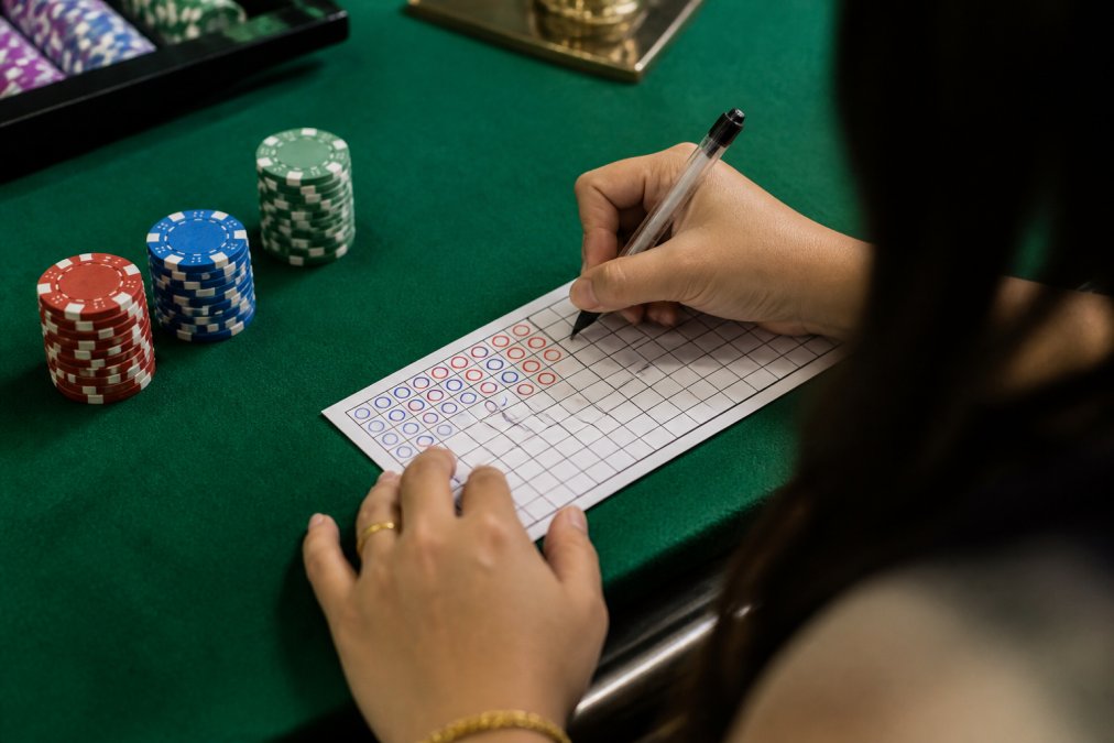Close-up of a player's hands marking a baccarat scorecard with a pen at a felt table, red and blue circles visible on the grid, chips stacked nearby
