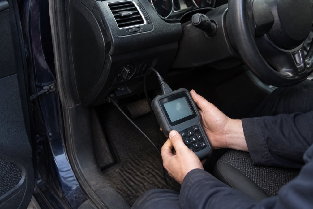 Mechanic's hands holding an OBD2 diagnostic scanner connected to a vehicle's diagnostic port, with the scanner screen displaying fault codes and live data in a well-lit garage workshop
