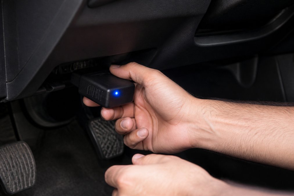 Close-up of hands plugging a Bluetooth OBD-II scanner into the port under a car dashboard, with the scanner's LED light visible