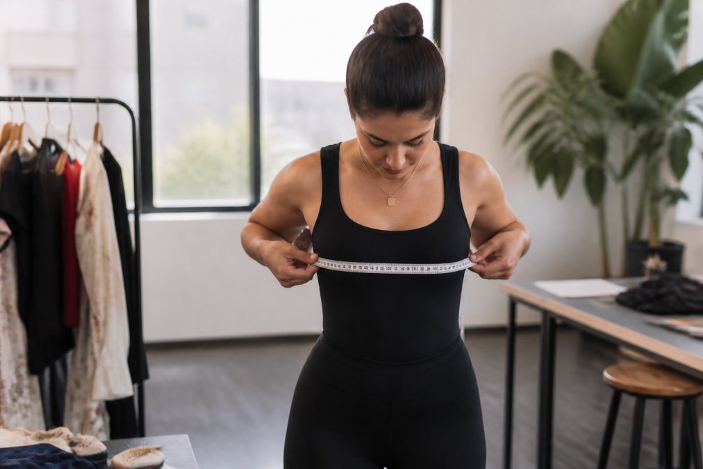Woman demonstrating proper body measurement technique, holding a flexible measuring tape around her bust with tape parallel to the floor, standing in natural lighting with arms relaxed at sides