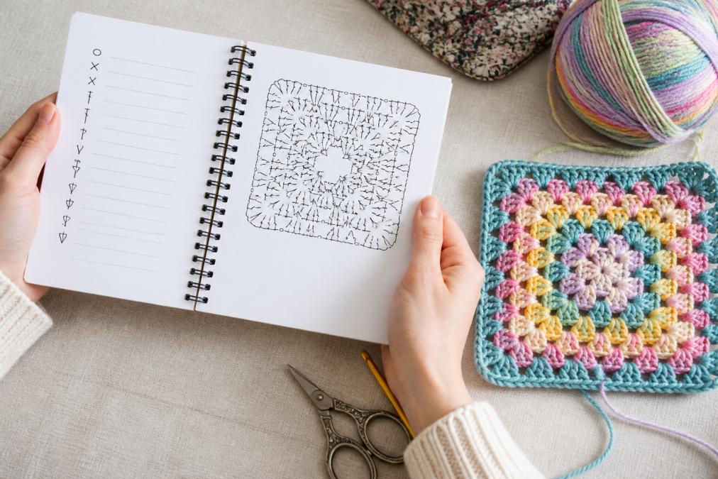 A pair of hands holding open a spiral-bound crochet pattern booklet showing a black-and-white symbol chart page, with a partially finished colorful granny square project and a ball of multicolored yarn resting on a light linen surface nearby