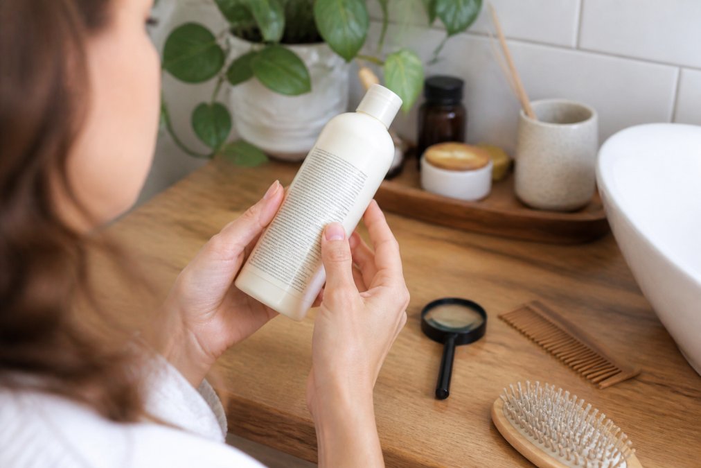 Close-up of a woman's hands holding a haircare bottle in natural light, reading the ingredient list on the back label with a magnifying glass nearby on a wooden bathroom counter
