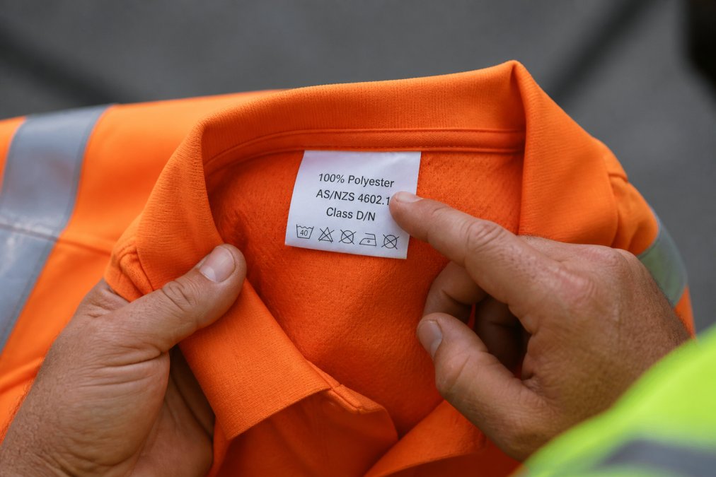 Close-up of a worker's hands holding the inside collar label of an orange hi vis shirt, pointing to the printed AS/NZS 4602.1 text and the row of care symbols beneath it, with the reflective tape visible in the background