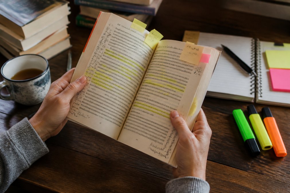 Person's hands holding an open book with handwritten notes, highlighter marks, and sticky notes visible on the pages, with a cup of tea on a wooden desk