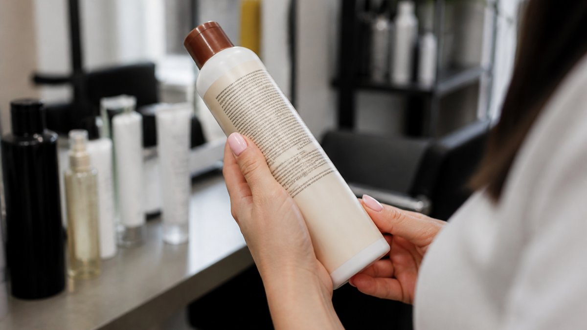 Close-up of hands holding a professional hair product bottle, turning it to read the ingredient list on the back label in a bright salon setting
