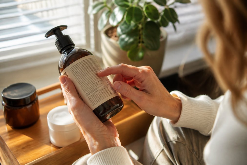 Close-up of a person's hands holding a skincare product bottle, carefully examining the ingredient list printed on the back label under warm natural window light